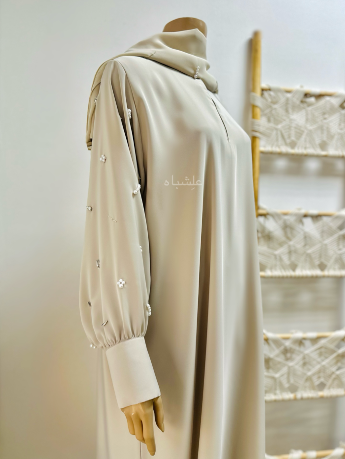 The Cream Nida Premium Fabric Abaya by Alishbah features a luxurious design with intricate embellishments of clip stones, pearls, and tube beads. Made from high-quality Nida fabric, the abaya drapes elegantly with a loose silhouette, ideal for both formal and casual occasions. Its neutral cream color allows for versatile styling, blending tradition with modern sophistication.