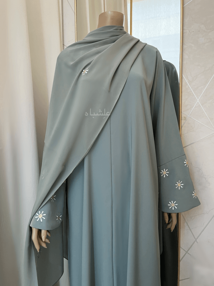 Abaya in green sage featuring premium Nida fabric with delicate floral embroidery on the overlapped sleeves and sheila. The Green Sage Nida Premium Abaya by Alishbah offers a blend of luxury, sophistication, and timeless elegance