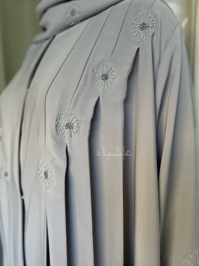 Alishbah Eternal Petals of Grace Nida Premium Abaya with floral handwork. The abaya is crafted from luxurious Nida Premium fabric, featuring delicate floral handwork including tiny scallops and French knots that form elegant patterns along the cuffs and neckline. The ensemble is completed with a soft chiffon sheila (headscarf) that drapes gracefully, enhancing the sophisticated and timeless design. This abaya blends traditional handwork with contemporary elegance, showcasing high-quality craftsmanship and refined style.