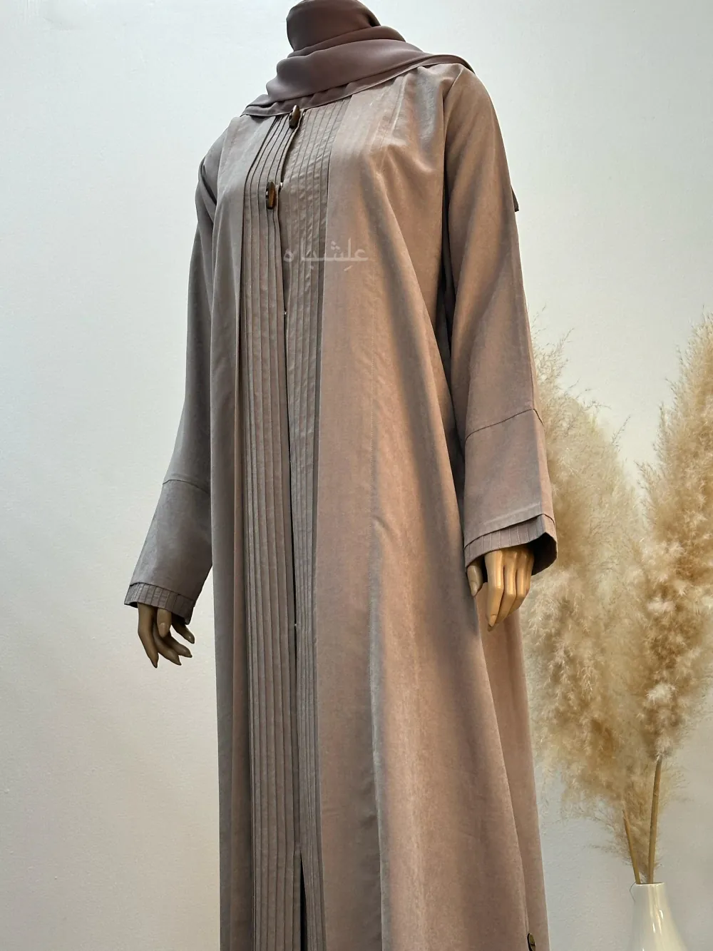 A beige abaya made from Hareer Magdhool fabric, featuring fine pleats down the front, two decorative buttons on the chest, and slightly flared sleeves, displayed on a mannequin.