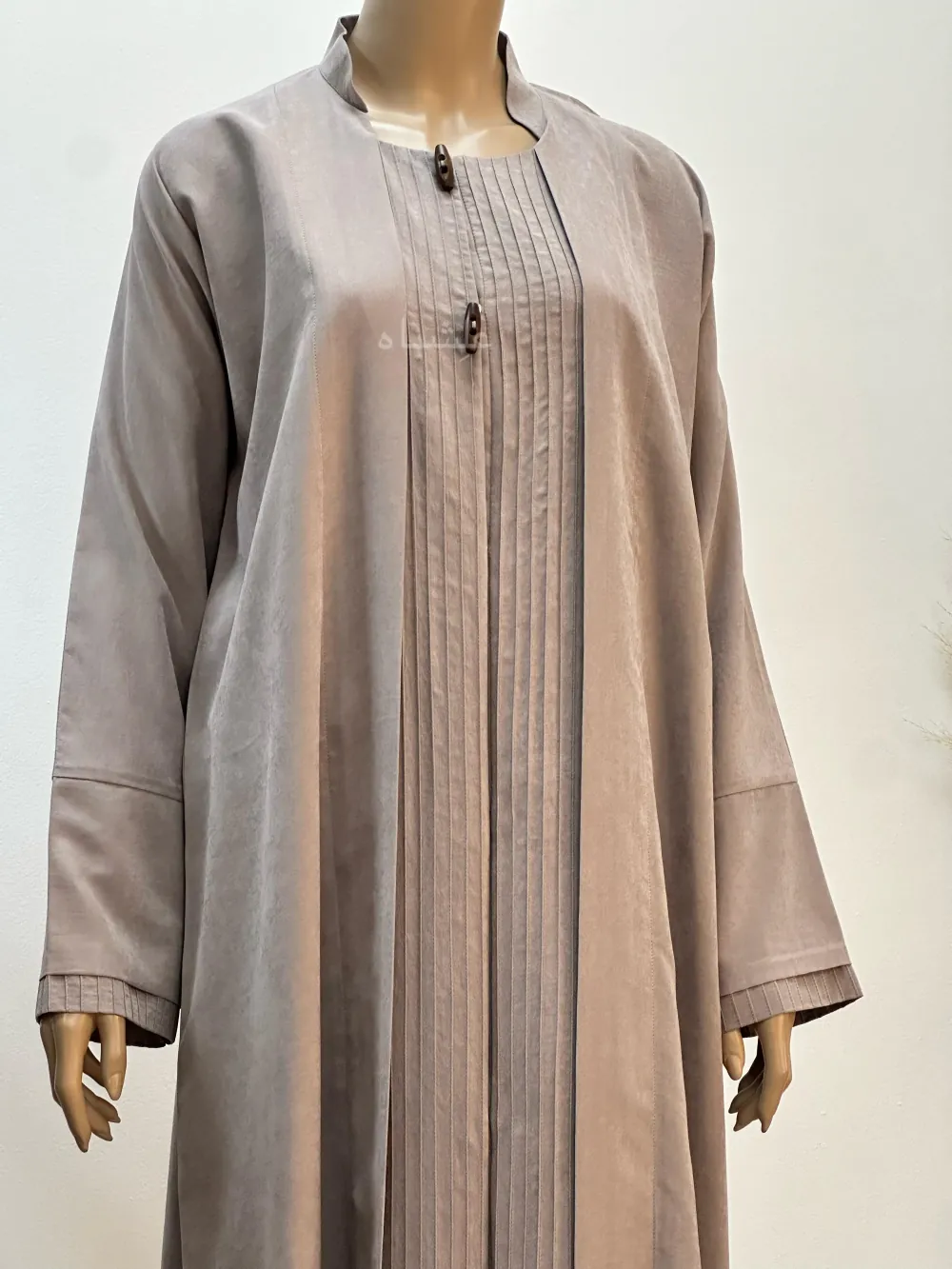 A beige abaya made from Hareer Magdhool fabric with fine pleats down the front, decorative buttons on the chest, and slightly flared sleeves, displayed on a mannequin.