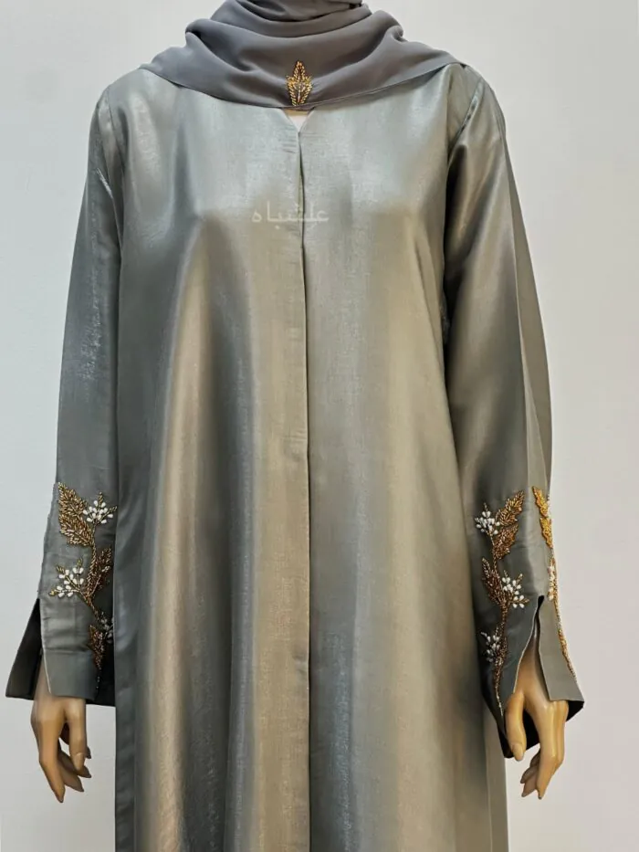 An elegant sage-colored abaya made from luxurious R.organza fabric, featuring intricate golden leaf embroidery on the sleeves with pearl-like bead accents. The abaya is complemented by a soft chiffon Sheila, draped gracefully, adding to the overall sophisticated look