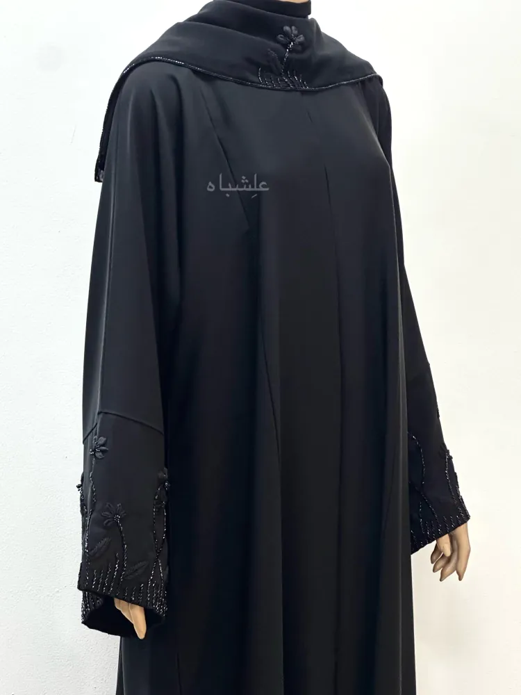 "Closeup of the Midnight Bloom abaya by Alishbah Abaya, with a chiffon sheila draped over the shoulder. The raised hands showcase intricate organza floral embroidery on the sleeves, highlighting the fine craftsmanship and smooth Nida fabric."