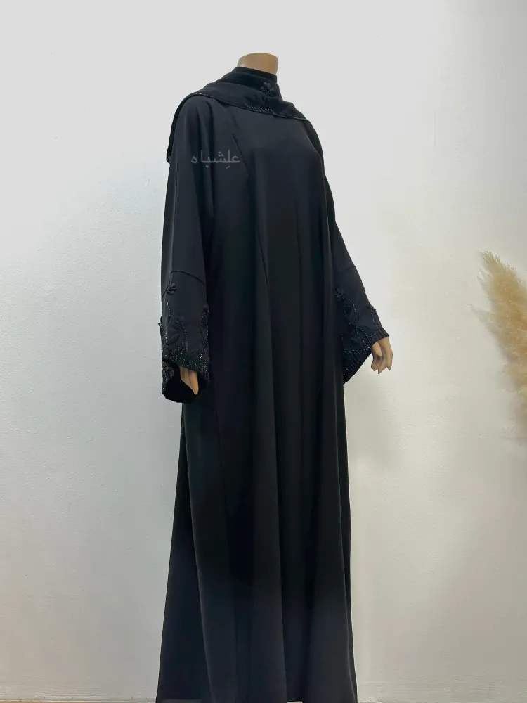 "Mannequin wearing the Midnight Bloom abaya by Alishbah Abaya, styled with a soft chiffon sheila. The figure is slightly turned to the side with raised hands, highlighting the organza floral embroidery on the sleeves and the seamless flow of the Nida fabric."